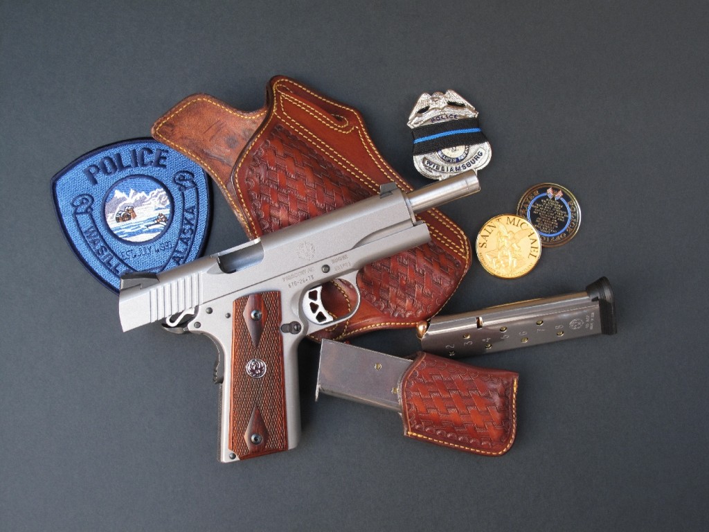 Le SR1911 culasse ouverte, accompagné de divers accessoires de la police américaine : un écusson brodé, un insigne barré par un ruban de deuil (thin blue line) et deux médailles (prayer coins) à l’effigie de Saint Michel, que les agents de police portent généralement sur eux, dans la poche ou à l’intérieur de leur képi, comme porte-bonheur.