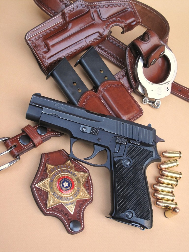 Le SIG-Sauer P220, présenté dans le contexte des forces de l’ordre aux USA, avec un badge de deputy (sheriff adjoint) de l’état du Texas, deux chargeurs de secours et une paire de menottes Smith & Wesson, dans un ensemble en cuir réalisé par la maison française La Sellerie du Thymerais.